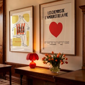 K&ouml;nnte beinhalten: Zwei gerahmte Poster an einer Wand, eines mit einem roten Herzen und dem Text "LES CHEMINS DE L'AMOUR ET DE LA VIE" und das andere mit einer Schwarzwei&szlig;illustration einer Person mit dem Text "CYRULIK SEWILSKI" und "opera poznani 1963". Eine rote Lampe mit einem blumenf&ouml;rmigen Schirm steht auf einem Holztisch mit einer Vase mit orangefarbenen Tulpen.