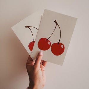 K&ouml;nnte beinhalten: Zwei rote Kirschen mit Stielen und Bl&auml;ttern, die auf eine wei&szlig;e Karte gemalt sind. Die Karte wird von einer Hand gehalten.