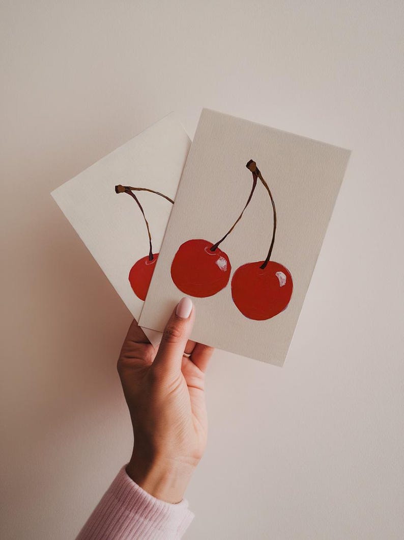 K&ouml;nnte beinhalten: Zwei rote Kirschen mit Stielen und Bl&auml;ttern, die auf eine wei&szlig;e Karte gemalt sind. Die Karte wird von einer Hand gehalten.