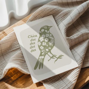 May include: A greeting card featuring a stylized green bird illustration filled with white floral patterns. The card has the text "Chica dee dee dee dee" in green. The card is on a wooden surface with a beige and white checkered cloth.