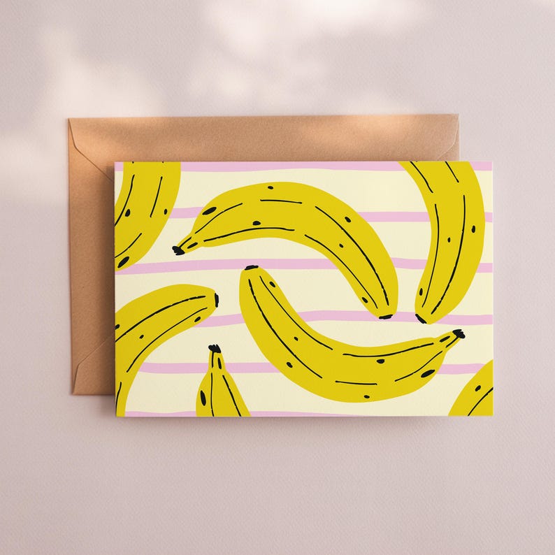 May include: Greeting card featuring a repeating pattern of yellow bananas with black spots on a cream background with pink stripes. The card is paired with a brown envelope, suitable for sending a note or message.