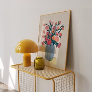 Könnte beinhalten: Eine gelbe pilzförmige Tischlampe und ein kleines grünes Glas stehen auf einem gelben Konsolentisch mit Drahtrahmen. Ein gerahmter Kunstdruck eines Blumenstraußes in einer blauen Vase lehnt an der Wand, wodurch eine stilvolle Wohnszene entsteht.