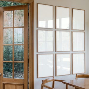 Op de afbeelding: Een witte muur met negen lege fotolijsten in een rasterpatroon. De lijsten zijn allemaal even groot en hebben een lichte houten afwerking. Links van de lijsten is een houten deur en voor de lijsten staat een houten tafel met stoelen.