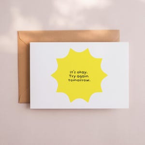 May include: A white greeting card with a yellow sunburst design and the text "It's okay. Try again tomorrow." is placed in front of a brown envelope. The card is on a light pink surface.