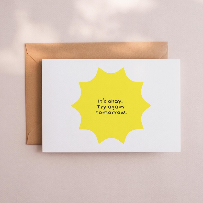 May include: A white greeting card with a yellow sunburst design and the text "It's okay. Try again tomorrow." is placed in front of a brown envelope. The card is on a light pink surface.
