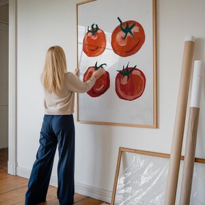Op de afbeelding: Een vrouw hangt een ingelijst schilderij van rode tomaten aan een witte muur. Het schilderij is in een eenvoudige houten lijst. De vrouw draagt een cr&egrave;mekleurige trui en een blauwe broek.