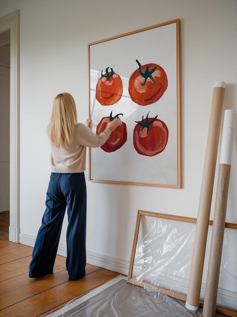 Op de afbeelding: Een vrouw hangt een ingelijst schilderij van rode tomaten aan een witte muur. Het schilderij is in een eenvoudige houten lijst. De vrouw draagt een cr&egrave;mekleurige trui en een blauwe broek.