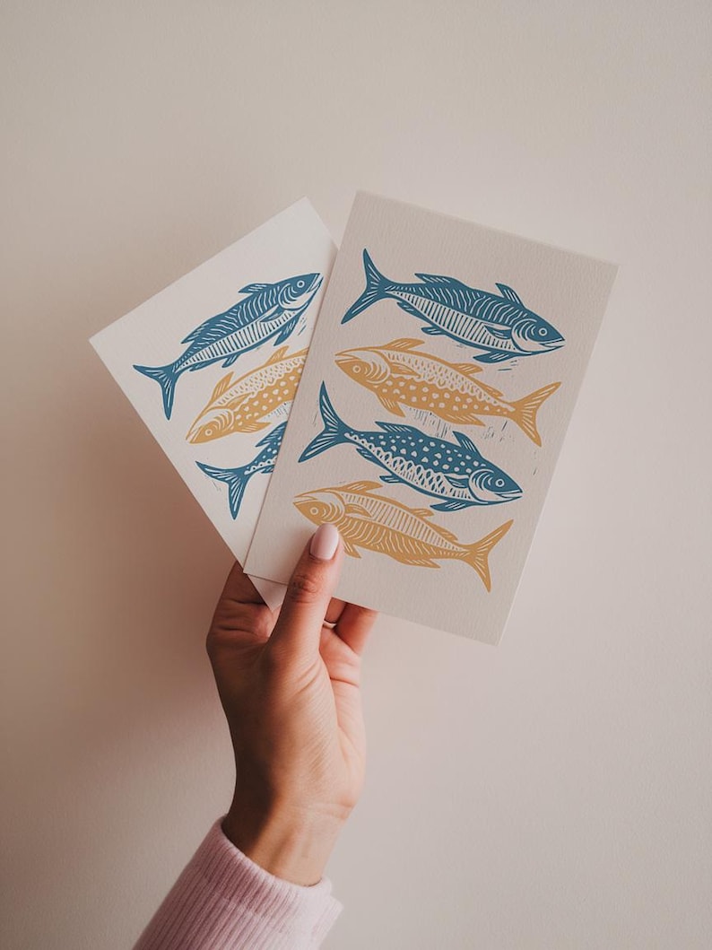 K&ouml;nnte beinhalten: Zwei Karten mit einem blauen und gelben Fischdruck. Die Fische sind stilisiert und haben ein klotziges Design. Die Karten werden in einer Hand gehalten.