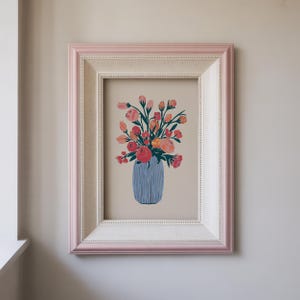Puede incluir: Una impresión artística enmarcada que muestra un jarrón azul lleno de flores rosas, rojas y naranjas. La obra de arte está enmarcada en un marco blanco con un borde rosa, ideal para la decoración del hogar.