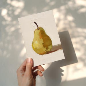 Puede incluir: Pintura al &oacute;leo de una pera amarilla con un tallo marr&oacute;n sobre un fondo blanco. La pera est&aacute; pintada con pinceladas visibles, y la obra de arte est&aacute; sostenida por una mano. La pintura est&aacute; iluminada por la luz del sol, proyectando una sombra.