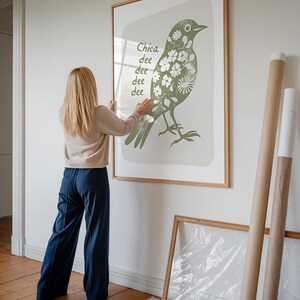 Op de afbeelding: Een vrouw hangt een ingelijst drukwerk van een groene vogel met witte bloemen aan een witte muur. De print heeft de tekst "Chica dee dee dee dee" erop.