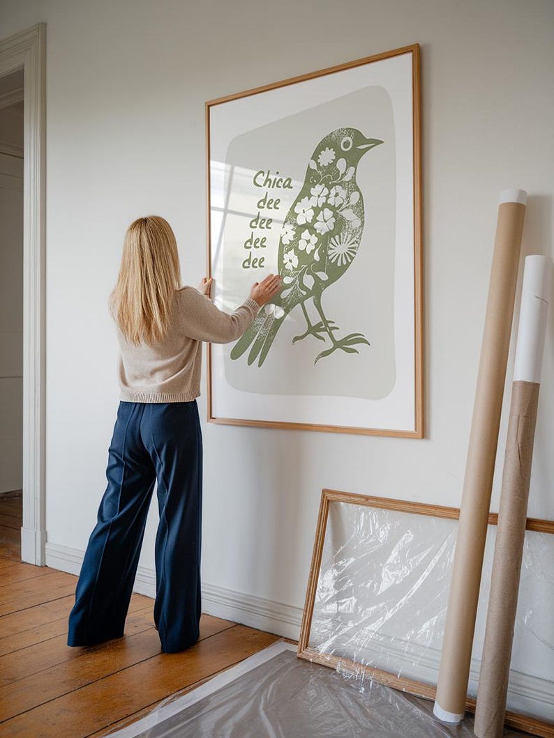 Op de afbeelding: Een vrouw hangt een ingelijst drukwerk van een groene vogel met witte bloemen aan een witte muur. De print heeft de tekst "Chica dee dee dee dee" erop.