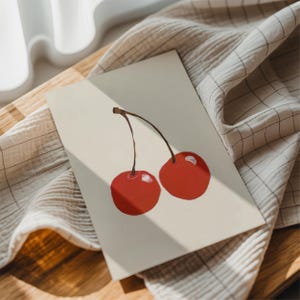 May include: A greeting card featuring a painting of two red cherries with brown stems. The card is on a light beige surface, with a beige and white checkered cloth in the background. The image is lit by natural light.