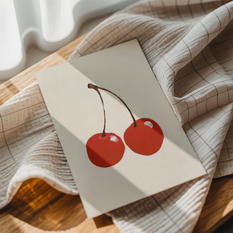 May include: A greeting card featuring a painting of two red cherries with brown stems. The card is on a light beige surface, with a beige and white checkered cloth in the background. The image is lit by natural light.