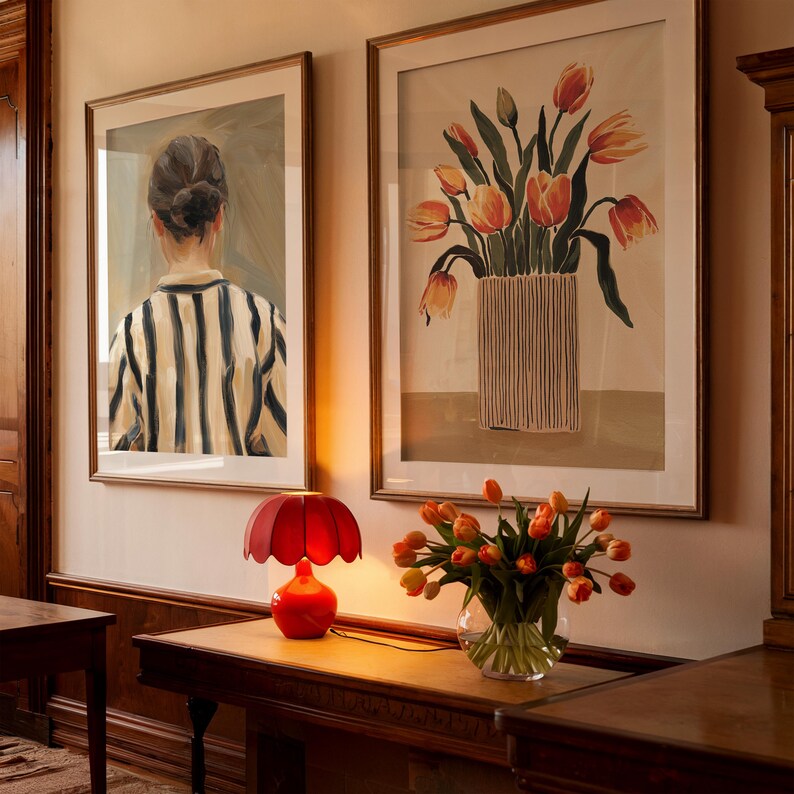 K&ouml;nnte beinhalten: Eine rote Lampe mit einem bl&uuml;tenf&ouml;rmigen Schirm steht auf einem Holztisch neben einer Vase mit orangefarbenen Tulpen. Zwei gerahmte Bilder h&auml;ngen an der Wand hinter dem Tisch. Ein Bild zeigt eine Frau mit dem R&uuml;cken zum Betrachter, die ein gestreiftes Hemd tr&auml;gt. Das andere Bild zeigt einen Strau&szlig; orangefarbener Tulpen in einer gestreiften Vase.