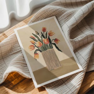 May include: A floral art print featuring a vase of orange and yellow tulips. The vase is beige with vertical lines. The print is on a white-bordered rectangular card, resting on a beige and white checkered cloth.