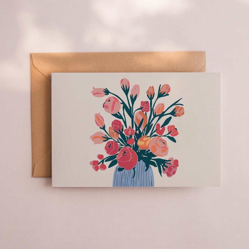 May include: Greeting card featuring a floral illustration. The artwork depicts a bouquet of pink, red, and orange flowers in a blue vase. The card is paired with a brown envelope, creating a soft, inviting aesthetic.