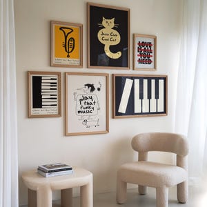 Puede incluir: Una pared blanca con siete carteles enmarcados, incluyendo un teclado de piano blanco y negro, una ilustración en blanco y negro de una persona tocando música, un cartel amarillo con una trompeta, un cartel negro con un gato y el texto "Jazz Club Cool Cat", un teclado de piano blanco y negro y un cartel con el texto "Love is all you need". Una silla blanca con textura esponjosa se encuentra frente a la pared.