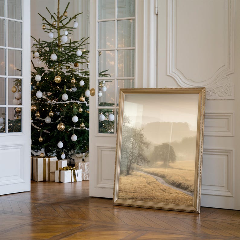 May include: A gold-framed picture of a foggy landscape with a path leading through a field. The picture is leaning against a white wall in a room with a Christmas tree decorated with white and gold ornaments.