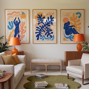 Op de afbeelding: Drie ingelijste abstracte kunstprints met een blauw, oranje en geel kleurenschema. De prints tonen gestileerde figuren en organische vormen.