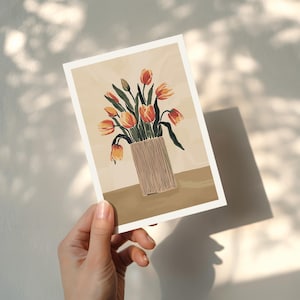 Puede incluir: Una impresi&oacute;n art&iacute;stica floral con un jarr&oacute;n de tulipanes naranjas y amarillos. Los tulipanes est&aacute;n en un jarr&oacute;n beige con l&iacute;neas verticales. El fondo es de color beige claro con una mesa marr&oacute;n. La impresi&oacute;n es sostenida por una mano.