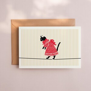 May include: Greeting card featuring a black cat wearing a red polka-dot dress and bow, walking a tightrope. The background has vertical beige and white stripes. A brown envelope is behind the card.