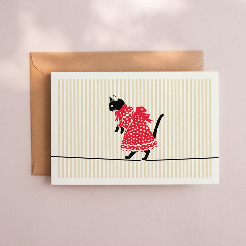 May include: Greeting card featuring a black cat wearing a red polka-dot dress and bow, walking a tightrope. The background has vertical beige and white stripes. A brown envelope is behind the card.