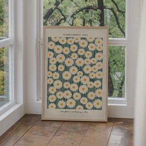 May include: A framed print with a white background featuring a pattern of white daisies with yellow centers on a teal green background. The text "FLOWER MARKET, ITALY" is at the top of the print and the text "Every flower is a soul blossoming in nature." is at the bottom.