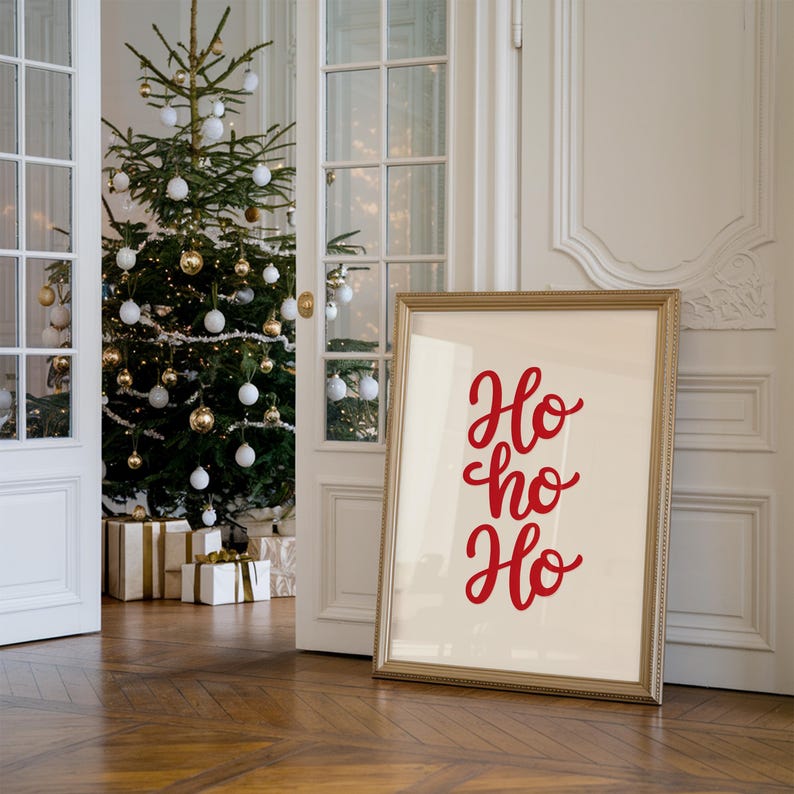 May include: A gold framed poster with the text "Ho ho ho" in red cursive lettering. The poster is leaning against a white wall in a room with a Christmas tree decorated with white and gold ornaments.