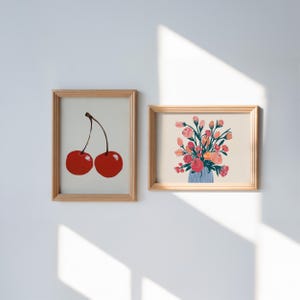 Könnte beinhalten: Zwei gerahmte Kunstdrucke. Einer zeigt zwei rote Kirschen mit einem braunen Stiel. Der andere Druck zeigt einen Strauß rosa und orangefarbener Blumen in einer blauen Vase. Beide sind in hellbraunen Holzrahmen.