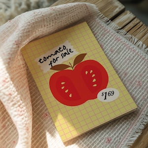 Puede incluir: Un cuaderno con un patrón de cuadrícula amarilla y una gran ilustración de tomate rojo. El texto "tomato for sale" está encima del tomate, y la etiqueta de precio dice "1,69 €".