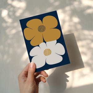 Puede incluir: Una tarjeta azul marino con dos flores estilizadas. Una es de color amarillo mostaza, la otra blanca, ambas con centros texturizados. La tarjeta se sostiene en una mano, con sombras proyectadas sobre la superficie.