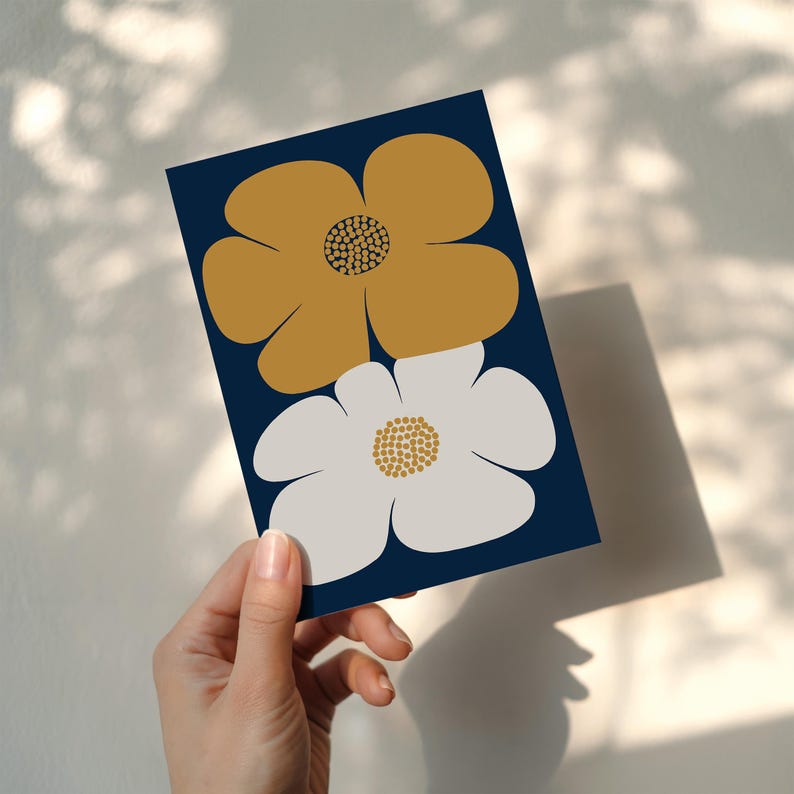 Puede incluir: Una tarjeta azul marino con dos flores estilizadas. Una es de color amarillo mostaza, la otra blanca, ambas con centros texturizados. La tarjeta se sostiene en una mano, con sombras proyectadas sobre la superficie.