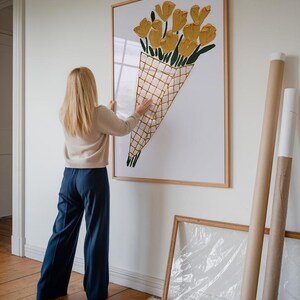 Op de afbeelding: Een vrouw hangt een ingelijst afdruk van een geel bloemenboeket aan een witte muur. Het boeket bevindt zich in een kegelvormige container met een geruit patroon.