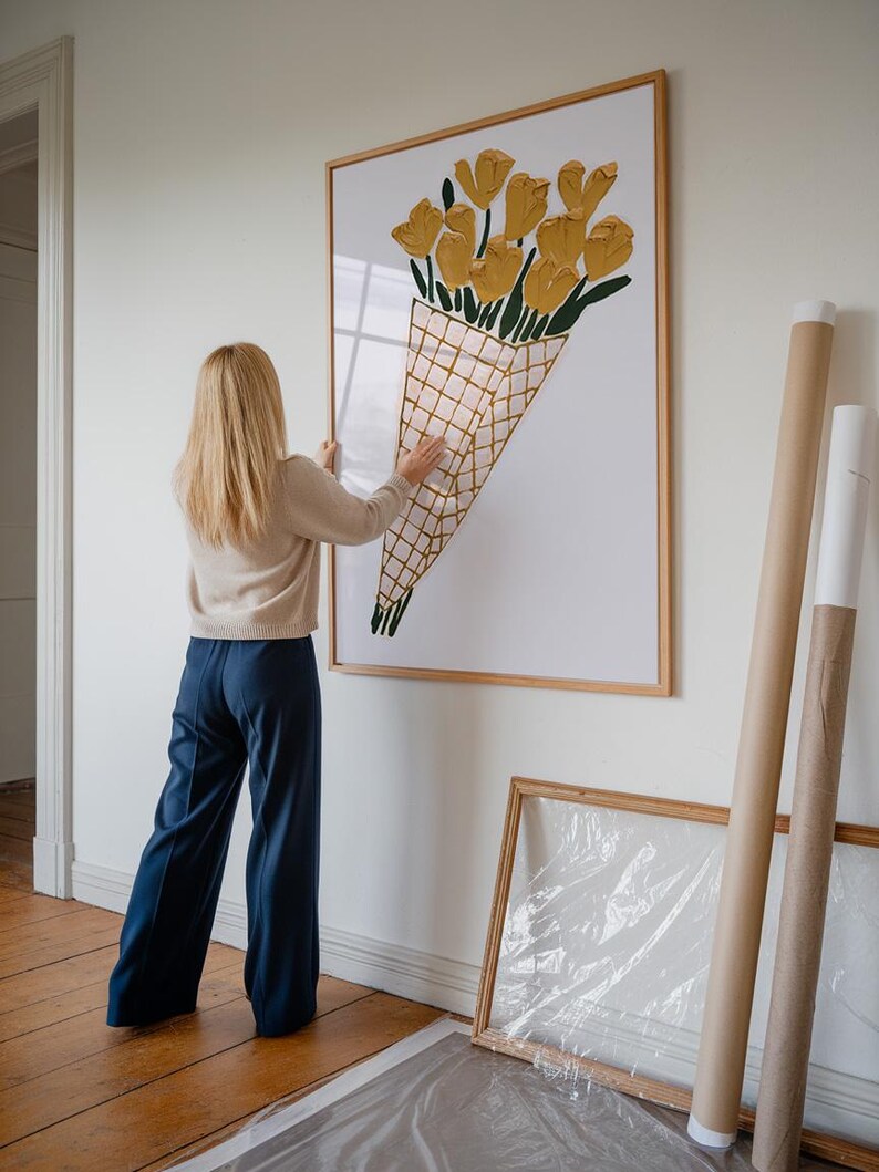 Op de afbeelding: Een vrouw hangt een ingelijst afdruk van een geel bloemenboeket aan een witte muur. Het boeket bevindt zich in een kegelvormige container met een geruit patroon.