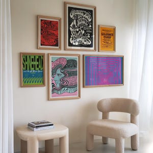 May include: A living room interior with a white wall featuring a collection of vintage concert posters. The posters are framed in wood and feature psychedelic designs and text. A beige armchair with a fluffy texture sits in front of the wall. A small beige table with rounded edges sits in front of the chair. A stack of books sits on the table.