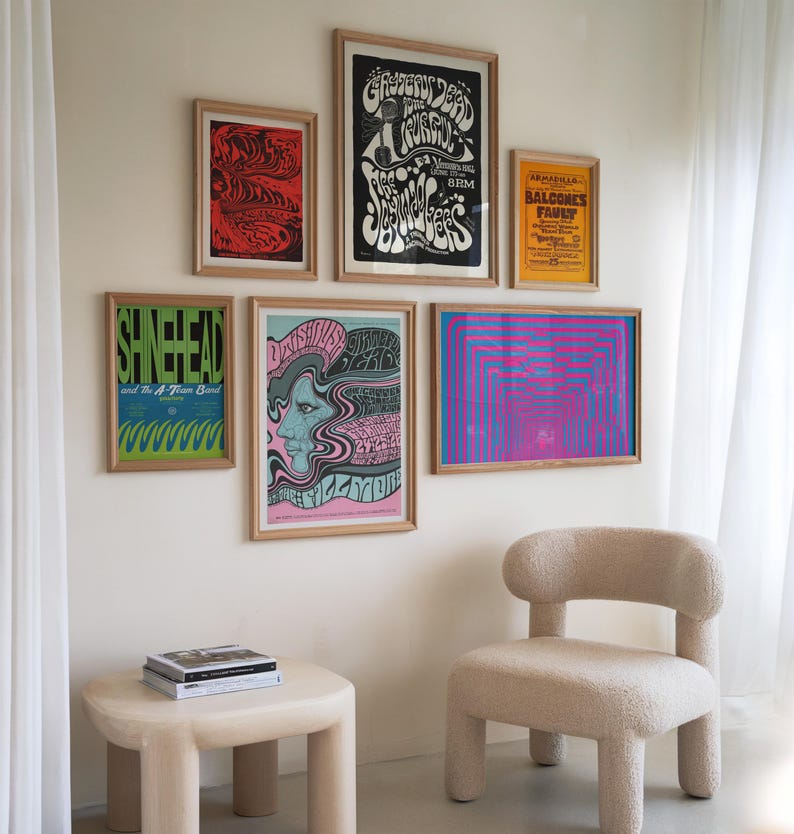 May include: A living room interior with a white wall featuring a collection of vintage concert posters. The posters are framed in wood and feature psychedelic designs and text. A beige armchair with a fluffy texture sits in front of the wall. A small beige table with rounded edges sits in front of the chair. A stack of books sits on the table.
