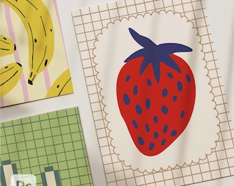 May include: Three colorful art prints featuring fruit illustrations. One print shows yellow bananas with black outlines on a background of pink stripes. Another print displays a red strawberry with blue seeds and a blue stem. The third print has a green grid pattern.