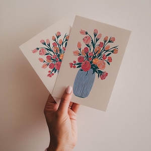 Puede incluir: Dos tarjetas con una ilustración floral de un ramo de flores rosas, rojas y naranjas en un jarrón azul. Las flores tienen tallos y hojas verdes.