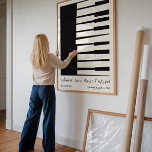 Op de afbeelding: Een ingelijste poster met een zwart-wit ontwerp van een pianoklavier. De poster heeft de tekst "Schoefer Jazz Music Festival" in zwarte handgeschreven letters. De tekst eronder luidt "in Central Park" en "Saturday, August 3, 1967".