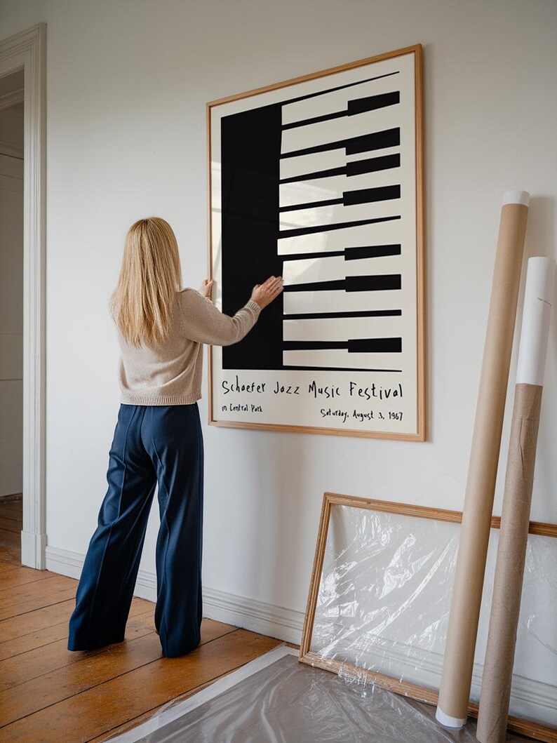 Op de afbeelding: Een ingelijste poster met een zwart-wit ontwerp van een pianoklavier. De poster heeft de tekst "Schoefer Jazz Music Festival" in zwarte handgeschreven letters. De tekst eronder luidt "in Central Park" en "Saturday, August 3, 1967".