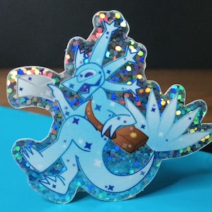 May include: A blue and white glitter sticker of a cartoon creature with a star-shaped head, holding a letter and a small bag. The creature has a long, flowing tail with a star-shaped tip.