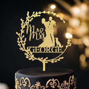 May include: A gold cake topper with the words "Mrs & Mrs" and "GEORGE" is on a black cake. The topper shows a couple and a dog inside a wreath. The cake has gold decorative patterns. The cake is a wedding cake.