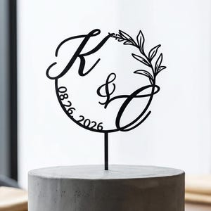 Op de afbeelding: Een zwarte caketopper met de initialen "K & C" en de datum "08.26.2026" in een cirkelvormig ontwerp met bladeren. De topper staat op een grijze cake, die op een zwarte plaat staat. De cake staat op een houten tafel.