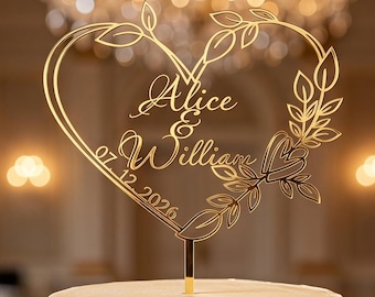 May include: A gold-colored cake topper shaped like a heart with leaf accents, featuring the names Alice & William and the date 07.12.2026 in script lettering. The topper is mounted on a thin stem and is positioned atop a two-tier white cake.