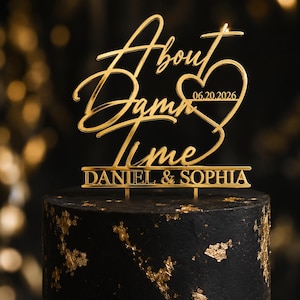 Könnte beinhalten: Ein goldfarbener Acryl-Tortentopper mit dem kursiven Text About Damn Time, einer Herzform, dem Datum 06.20.2026 und den Namen DANIEL & SOPHIA in Blockschrift, montiert auf zwei kleinen Stiften auf einer schwarzen Torte mit Blattgold.