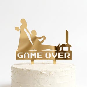 May include: A gold-colored acrylic cake topper featuring a silhouette of a person in a dress holding a bouquet standing behind a seated person playing a video game. The base of the topper displays the pixelated text GAME OVER in gold.