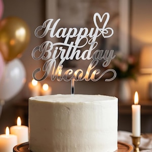Puede incluir: Un adorno para pastel de acrílico plateado con el texto "Happy Birthday Nicole" en letra cursiva y un icono de corazón en la parte superior. El adorno está insertado en un pastel redondo con glaseado blanco sobre una base de madera.