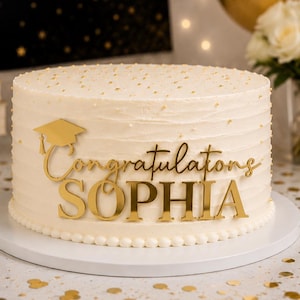 Op de afbeelding: Een ronde taart met wit glazuur, bestrooid met gouden spikkels en voorzien van een gouden acryl taarttopper met een afstudeerhoedje, de tekst Congratulations in cursief schrift en de naam SOPHIA in grote hoofdletters op de voorzijde.