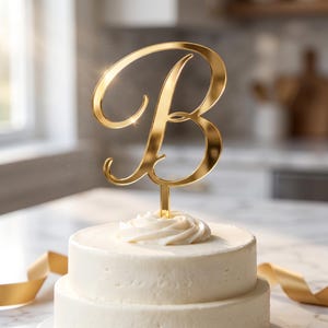 May include: A gold-colored cake topper featuring the letter B in a cursive script font. The topper has a reflective, metallic finish and is mounted on a thin stem, positioned upright in the center of a white frosted two-tiered round cake.